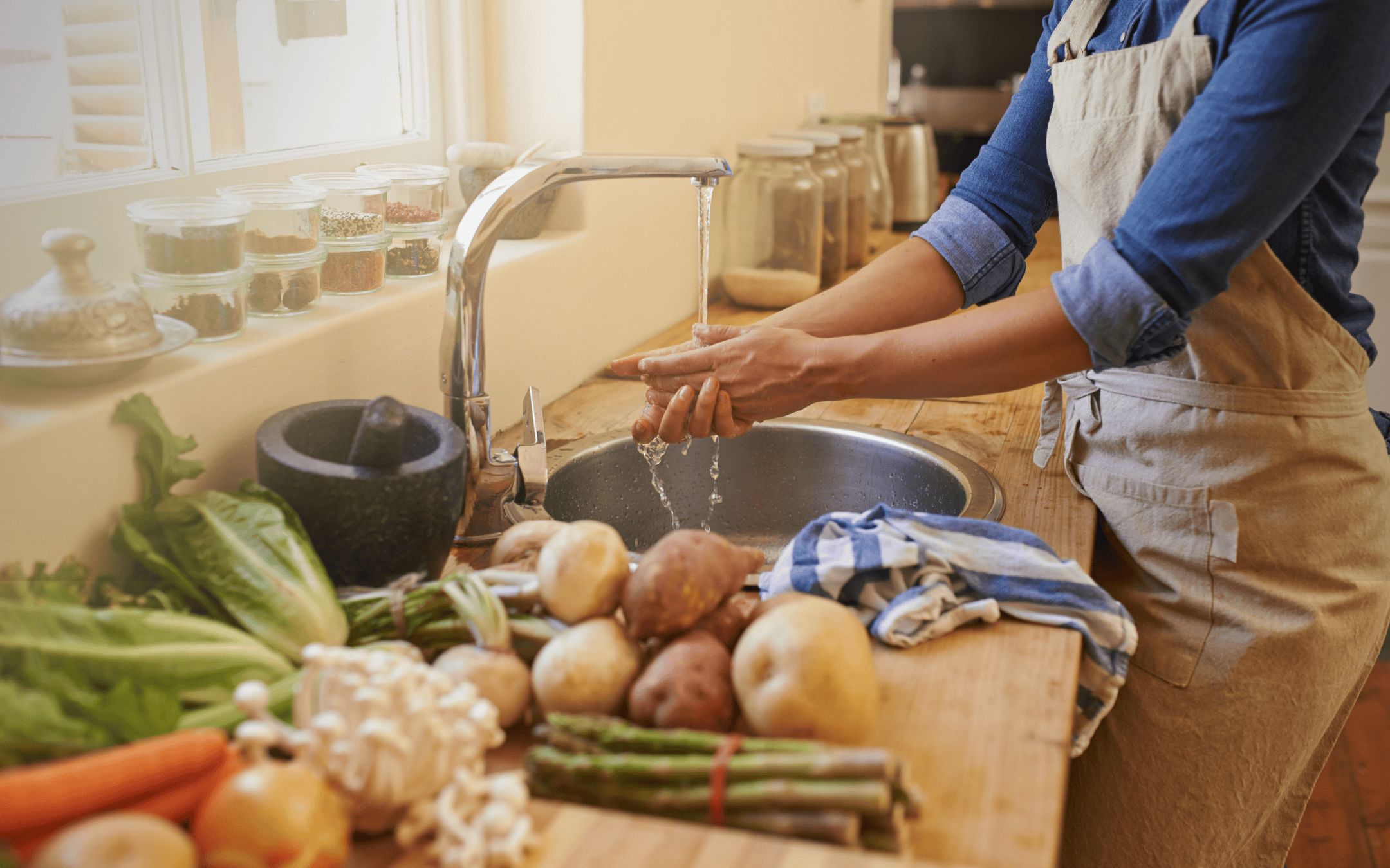 Person washing hands in a kitchen that’s full of green leafy vegetables, root crops, mushrooms, herbs, grains, and other healthy ingredients - symbolizing healthy and organic food choices to control autoimmune disease triggers.
