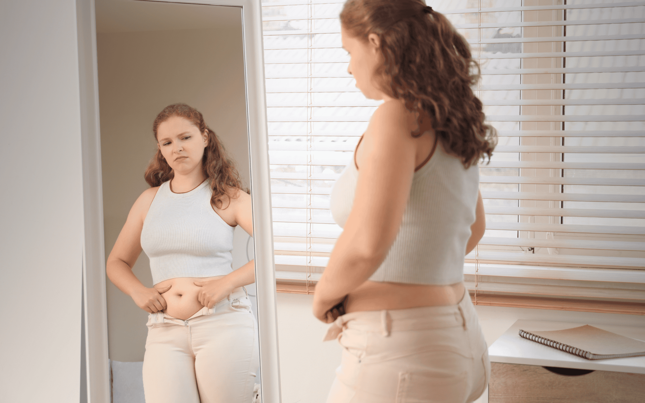 Woman looking at the mirror while clutching her lower belly, frustrated by tight unzipped jeans and weight gain, reflecting common thyroid symptoms often missed by "normal" lab results.