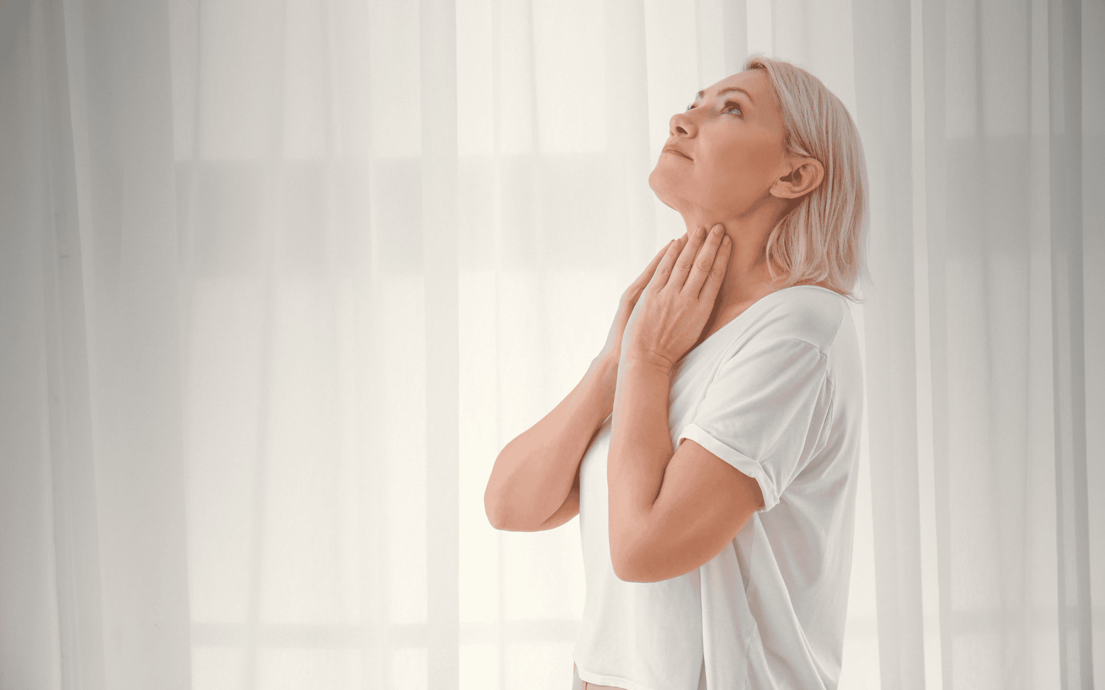 Woman in white gently touching her neck, looking up with a peaceful expression, symbolizing self-awareness and the search for answers in understanding thyroid health and autoimmune conditions.