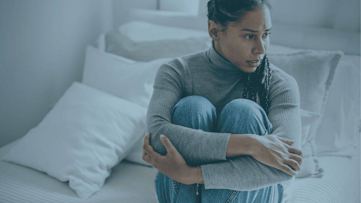 Woman sits on bed while hugging her knees with a worried expression on her face, illustrating anxiety possibly linked to low progesterone.